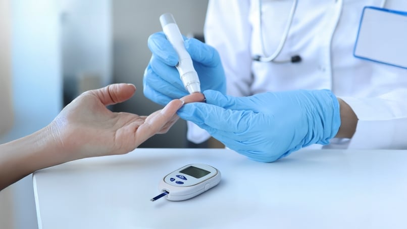 Doctor with gloves measuring blood glucose by piercing patient's finger with lancet and glucometer placed on table