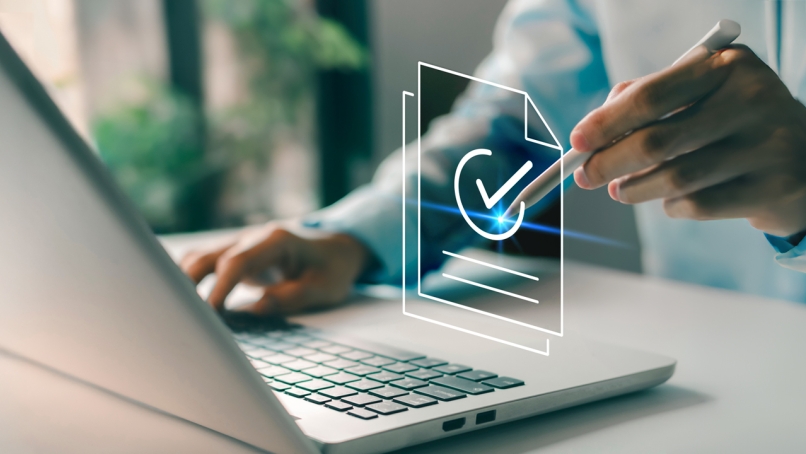 Person's hand pointing at compliant documents hologram with checkmark in front of a laptop