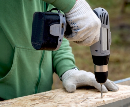 Person's wearing white utility gloves and drilling holes in a wooden board with a power drill.